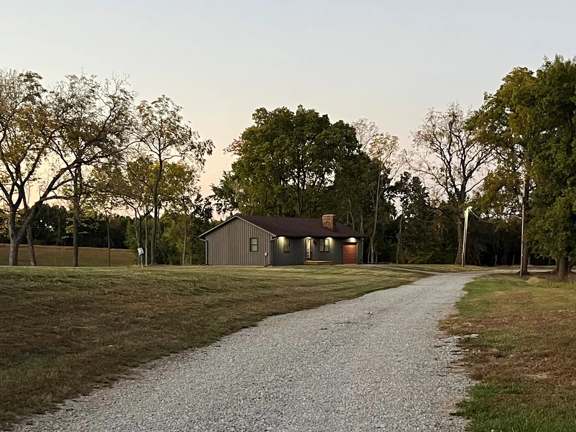 The farmhouse at dusk at the end of a gravel drive, surrounded by trees